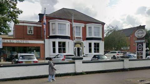 Exterior of the Pearl Hotel- a white and red building with a Tavan signboard outside in the car park- and an England flag hoisted next to the building- and a member of the public- a woman wearing a grey coat and blue jeans walking in front of it.