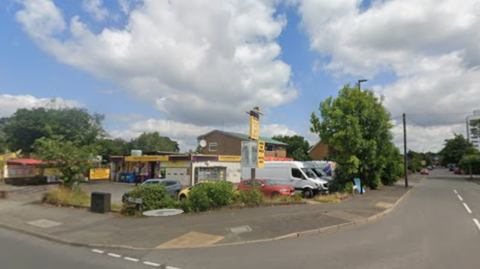 Street-corner view of a suburban road junction on a bright day, with a small commercial building and forecourt on the left, several parked vehicles including white vans and cars, trees and shrubs along the roadside, and a wide blue sky filled with large white clouds. A two-lane road extends into the distance past residential and commercial properties.