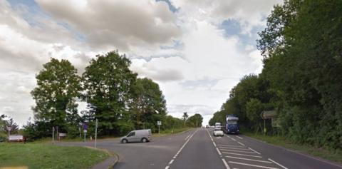 The A5 at a junction with a side road, looking south. Traffic from the south has a filter lane for those using the right-hand turn into the side road. A van has just entered the side road. There are tall trees on the south side of the junction. A signpost is on the right. A blue lorry is heading north while a white car has just taken the filter lane. There are clouds in the sky.