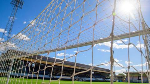 Close up of the netting of one goal at the Abbey Stadium, Cambridge, with the football pitch, a grandstand and floodlights behind. 