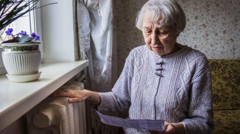 An elderly lady in her front room looks anxiously at a paper bill in her hand, with her other hand resting on a radiator. She has silver hair and is wearing a knitted jumper. 