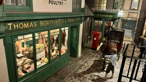 The Victorian village, with a cobbled street, a shoeless boy leading a horse through the street in front of a butchers shop