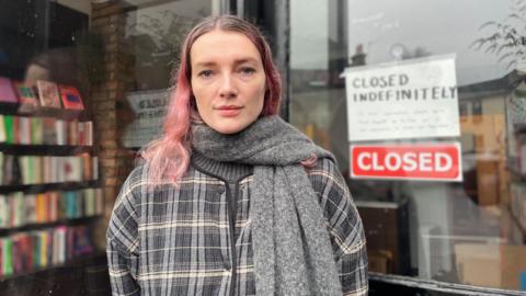 A woman with long pink hair and a grey striped jacket stands outside a shop. Behind the glass are rows of books. A sign on the door reads 'Closed Indefinitely'