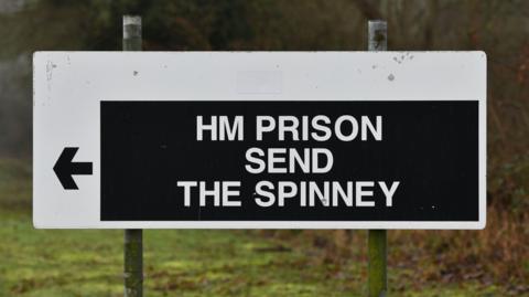 A sign reading HM PRISON SEND THEN SPINNEY written in white lettering with a black arrow pointing to the left