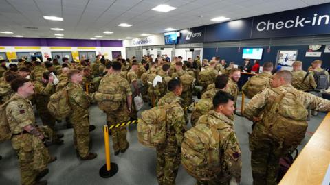 Lines of soldiers at the check-in desks at the ferry port. They are all wearing camoflauge clothing and have backpacks.