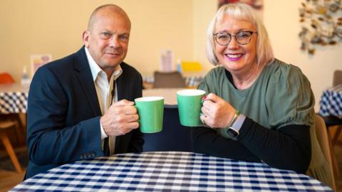 John Kinght and Mary Doyle sitting at a round table with a black and white chequered tablecloth in a cafe holding up matching green mugs. He is wearing a dark blue jacket over a white shirt and has closely cropped fair hair, while she has shoulder length blonde hair and it wearing glasses and a green and black top.