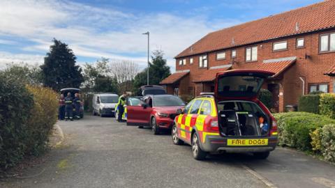 An emergency vehicle with "Fire Dogs" on it is parked up on the pavement outside a row of houses. Fire crews surround another vehicle and another emergency vehicle is tucked behind a parked car.