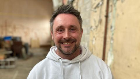 A man with brown hair and a short beard is wearing a white hoodie. He is standing in a room which appears to be mid-way through a renovation project. He is smiling for the camera.