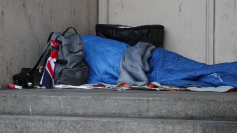 A person is lying in a sleeping bag on a blanket at the top of a set of stone steps. They also have a coat thrown over them. There is a small backpack near their head and a pack of cigarettes and a larger case on their other side of them.