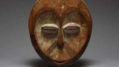 A mask depicting a stylised version of a human face - there is a small smile, narrow eyes and dots pressed into surface which go in a horseshoe shape where eyebrows would be and are also under the nose.