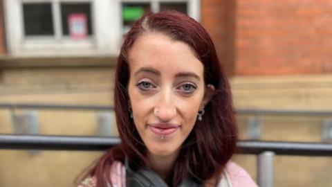 A girl with red hair and facial piercings looking at the camera, with a pink jumper on and head phones on 