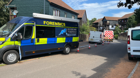 View of a cul-de-sac that has chalet-style houses. A large police forensics van is parked up on the pavement, three other vans parked up too. Small cones with police tape attached are up. It is a sunny day.