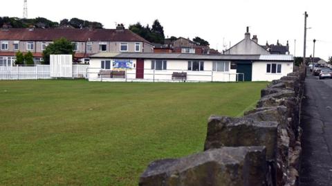 A single storey white building with a large green cricket pitch in front of it and a low stone wall on the right next to a road