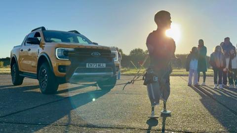 A large yellow four-wheel drive vehicle is on the left, attached by a cord and ropes to triple amputee Mark Ormrod, who stands in the sunlight.