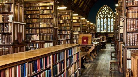Ancient looking wood-panelled library with rows and rows of books and stained glass window in background. 