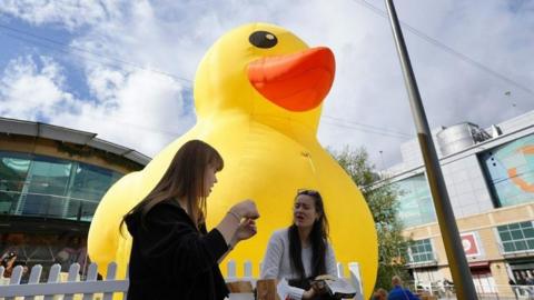 A large rubber duck is placed behind two women chatting on a bench while eating food. There are buildings in the background that look like shops. 