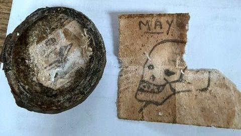 A picture of part of what Alex discovered. A round object is on the left of the picture whilst a note, hand-drawn with a skull, teeth and dagger in his head can be seen on the right.