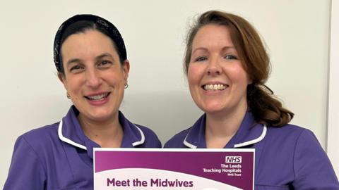 Two midwives in purple uniforms stand against a plain white wall, holding a poster about a new support session for expectant and new parents. 