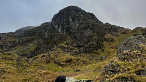 Priest's Hole at Dove Crag in the Lake District. It is a mossy, black rock face that has broken, grassy rocky ground leading up to it.