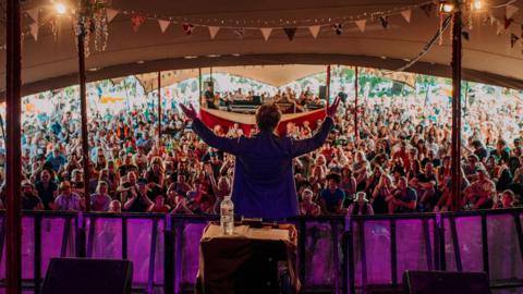 A Shindig festival tent filled with people as far as the eye can see is pictured from the back of the stage. The performer is standing with their back to the camera, looking at the crowd with their hands up. 