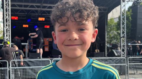 A young boy is stood outside smiling in front of a stage dimly lit behind him