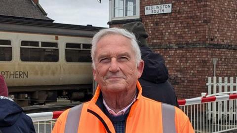 Glen Sanderson smiling into the camera. He has white hair and is wearing an orange hi-vis coat over a jumper and shirt. Behind him is the railway station which has a brick signalling tower on the right. A worn-looking grey Northern train is at the station. Two people stand behind him facing away from the camera in hats and coats. The railway barrier is down.