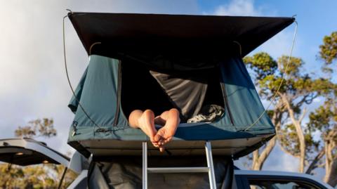 A green tent box on top of a car, with a pair of feet sticking out from it