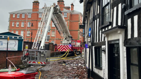 A black and white timbered building with a pile of rubble and a fire engine in the street outside