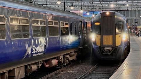 ScotRail carriages and a ScotRail locomotive with headlights on are waiting on two separate railway lines in a terminus station. People can been seen in the distance under a sign for platform 4.