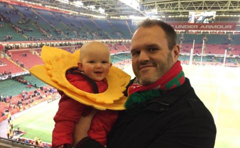 Rugby fan Sam watching a game with his young child - the child is wearing a daffodil hat. He is turning away from the stadium and looking at the camera holding his child. The pitch is behind him