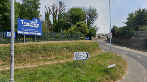 A blue sign with the word 'Trenance' on it in white writing. There is smaller text below it and an arrow pointing up to the entrance. There is a smaller white direction sign near the road on a grassy area. 
