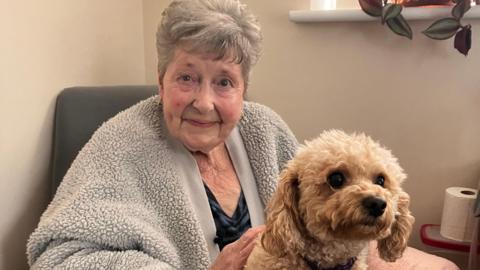 Pat Ellis at home with her dog she's wearing a grey fleece top over a navy blouse. She has grey short hair and is smiling at the camera. Her small brown dog is on her lap.