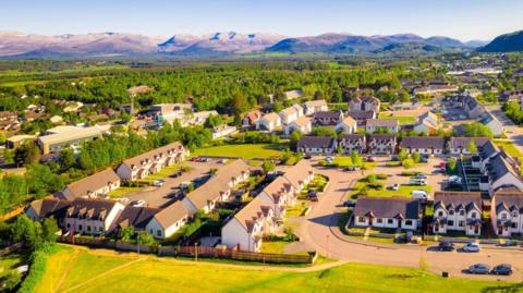 An aerial view across part of Aviemore on a sunny day. There are rows of houses and large areas of trees. There are hills and mountains on the horizon.