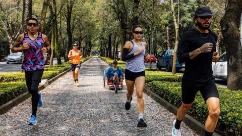 Five people running on a tree-lined paved path with roads either side. The dappled sunlight is shining down.