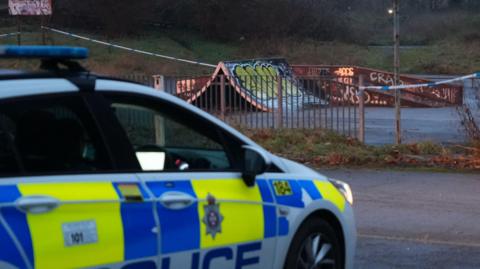 A police car parked in front of an old skatepark with graffiti all over it. There are railings around the skatepark and police tape.
