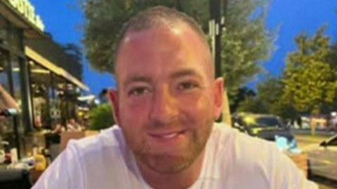 Stephen Reardon is smiling at the camera. He has short black hair and a short brown beard. He is wearing a white T-shirt.