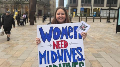 A woman with long dark hair and a smiling face holds a placard which reads "Women need midwives" the rest of the placard is not in view in this picture. She is standing on a square with pedestrians in the distance.