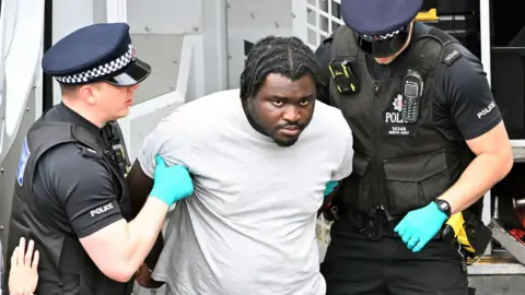 Anthony Esan, dressed in a grey tracksuit, with his arms being held behind his back by male uniformed police officers