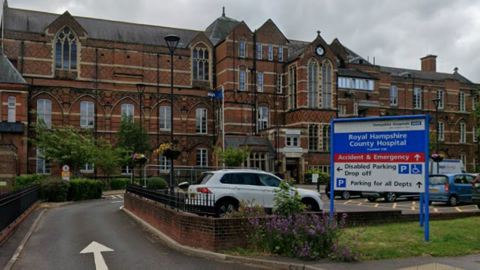 The older part of Royal Hampshire County Hospital is an ornate brick building of three or four storeys with large windows and a modern hospital sign in front pointing the way to departments.
