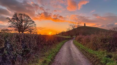 Sunrise on a winter's day in the countryside, looking along a deserted lane bounded by hedgerows and trees towards Glastonbury Tor. The Tor is silhouetted against vibrant oranges of the dawn sky and clouds.