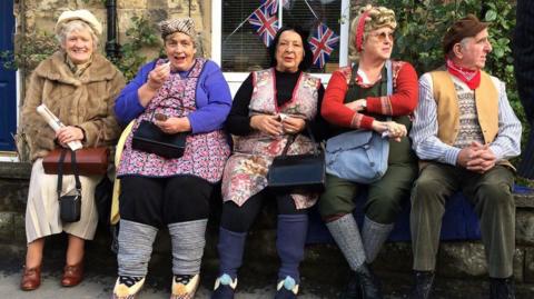 Four women and one man sit next to each other along a stone wall, looking cheerful. All of them are wearing 1940s style clothing, such as fur coats, aprons, and waistcoats. There is Union Jack bunting in a window behind them.