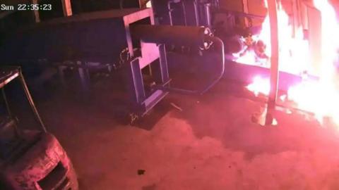 A still from CCTV footage shows a piece of industrial machinery on fire. The fire is in the right hand corner of the image. There is a piece of machinery in the centre of the image and a vehicle on the left, neither of which are on fire.