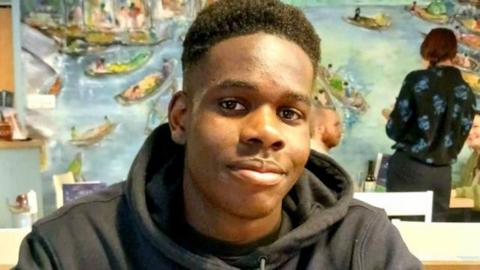 Shayne Hambakachere. He has short black hair and is pictured wearing a black hoodie. He is looking directly at the camera and smiling.