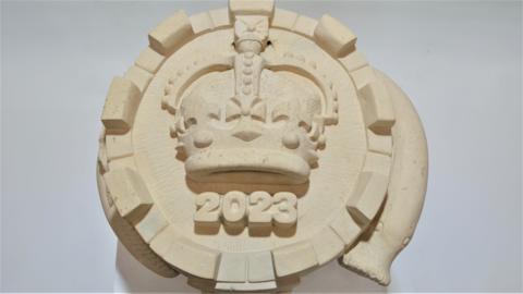 A circular piece of stone with a monarch's crown in the middle, 2023 written underneath it and a lamprey snaking around its sides.