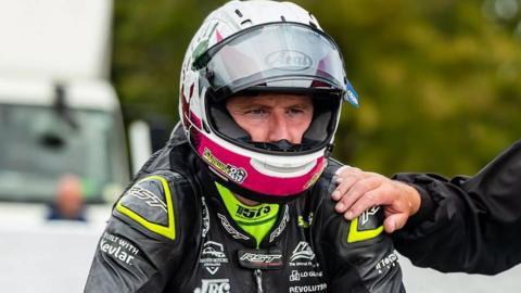 Sam Naughton in his racing leathers and helmet on the start line with a hand on his shoulder.