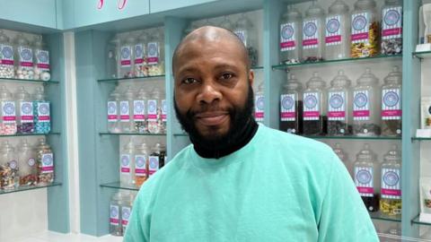 Leion Dillon stood inside his sweets store in Gallowtree Gate, Leicester. He is wearing a green shirt and behind him are traditional tubs of sweets