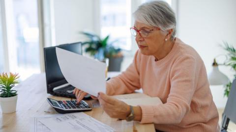 Older woman sits at a desk at home holding a bill with her other hand on a calculator and a laptop in the background.