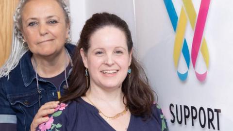 Steffi Colbert and Nat are both looking at the camera and smiling. Steffi stands to the left of the image, just behind Nat. She has grey dreadlocks in a half up half down hairstyle and wears a dark blue denim shirt with a deep purple cotton round neck t shirt underneath, her hand is resting on Nat's shoulder. 