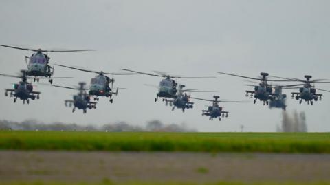 Dozens of helicopters lift off from an airfield on a grey day. They are in a close formation as they fly.