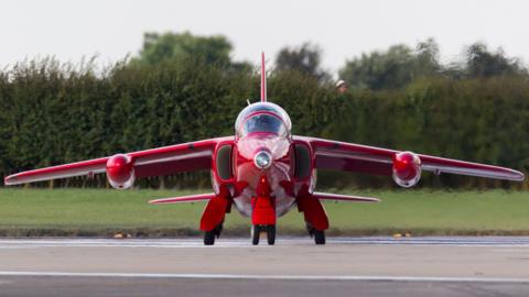 Front view of a Red Arrows jet taxiing at RAF Waddington. There are trees in the background.